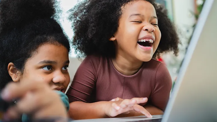 Two young African American girls laughing and playing on a computer.